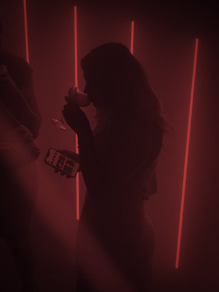 Silhouet van een vrouw die een cocktail drinkt terwijl ze een telefoon vasthoudt, met verticale lichtstralen op de achtergrond