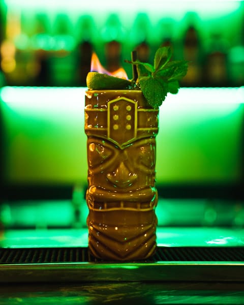 Tiki-cocktail geserveerd in een keramisch glas met een vlam, munt en limoen, op een bar met groene achtergrondverlichting.