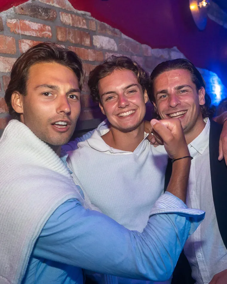 Drie lachende mannen tijdens een cocktailavond in club Roto, met promotekst 'Dinsdag' in beeld